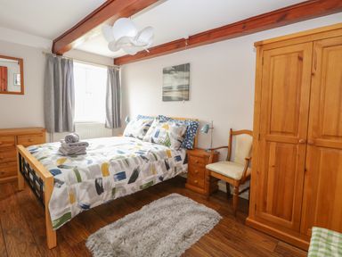 A bedroom with a bed dressed in patterned bedding a wooden wardrobe a wooden chest of drawers a wooden chair and a window with gray curtains at Compass Cottage in Bodham near Sheringham