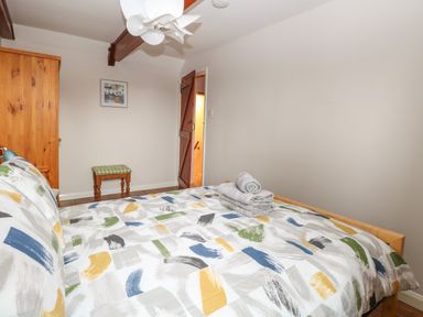 A bedroom with a bed covered in a patterned duvet a wooden wardrobe a small stool and a door open to a bathroom at Compass Cottage in Bodham near Sheringham