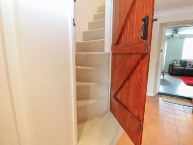 A staircase with beige carpet next to a wooden door and a living room with a black sofa and red rug at Compass Cottage in Bodham near Sheringham