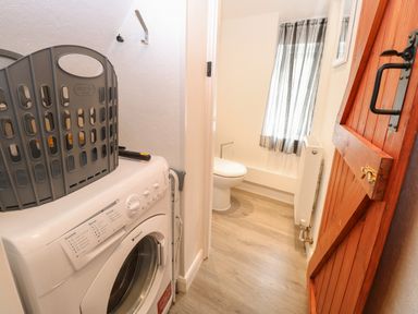 A laundry area with a washing machine and a gray basket next to a small toilet room with a wooden door and a window with sheer curtains at Compass Cottage in Bodham near Sheringham