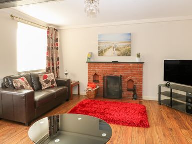 A living room with a brown leather sofa red and beige cushions a red rug brick fireplace and a TV on a glass stand at Compass Cottage in Bodham near Sheringham