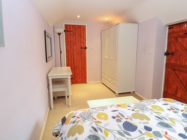 A bedroom with a bed covered in a floral duvet a white wardrobe a white desk with a chair a floor lamp and two wooden doors at Compass Cottage in Bodham near Sheringham