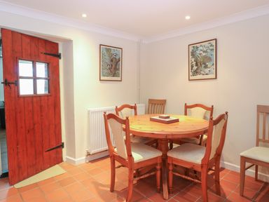 A dining area with a round wooden table and five chairs near a red wooden door at Compass Cottage in Bodham near Sheringham