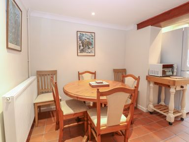 A dining area with a round wooden table and five chairs a microwave on a wooden cart and framed pictures on walls at Compass Cottage in Bodham near Sheringham