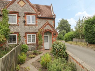 A brick and pebble stone cottage with white framed windows and a tiled roof beside a narrow road at Compass Cottage in Bodham near Sheringham