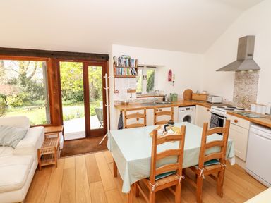 A kitchen with wooden chairs and table with a light green tablecloth and a white sofa near glass doors at Puffin Cottage in Hawkchurch