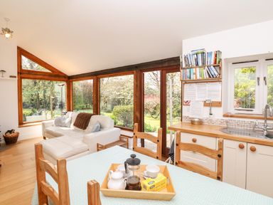 A living room with a white sofa and large wooden framed windows next to a kitchen area with a wooden countertop and a dining table in front with a tray containing a teapot and cups at Puffin Cottage in Hawkchurch