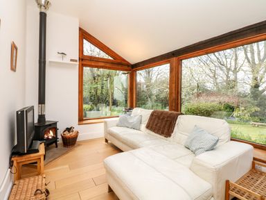 A living room with a white sofa, wood burning stove, television on a wooden stand, and large windows showing a garden at Puffin Cottage in Hawkchurch