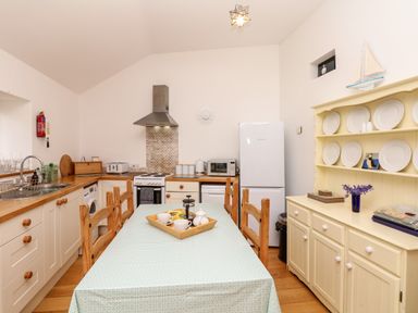 A kitchen with a dining table covered by a tablecloth and wooden chairs a white refrigerator a stove a wooden cabinet with plates and a decorative boat model at Puffin Cottage in Hawkchurch
