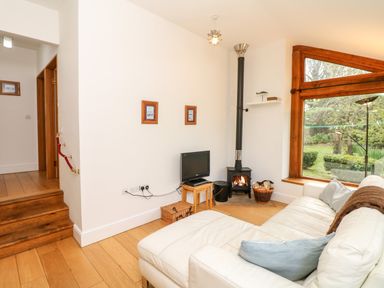 A living room with a white sofa and pillows a small television on a wooden stand a wood-burning stove and a large window showing a garden at Puffin Cottage in Hawkchurch