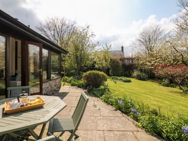 A patio with table and chairs next to a garden with trees and flowers at Puffin Cottage in Hawkchurch