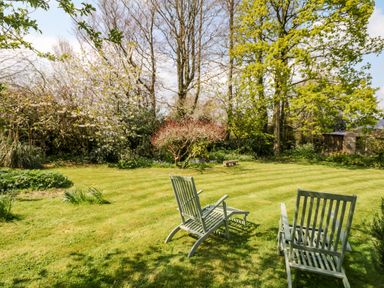 A garden with two wooden chairs on grass surrounded by trees and shrubs at Puffin Cottage in Hawkchurch