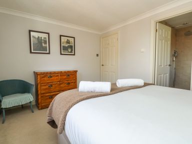 A bedroom with a bed holding two rolled towels a wooden chest of drawers a green chair and an open door to a bathroom at Gabrianne in Burton Bradstock