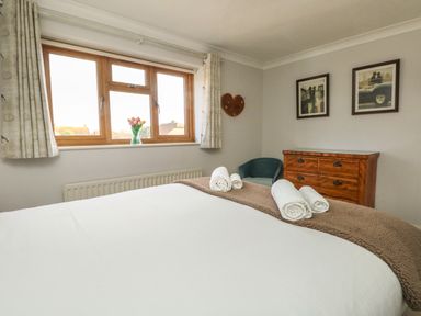 A bedroom with a bed, rolled towels, a wooden dresser, a blue chair, framed pictures, and a window with curtains at Gabrianne in Burton Bradstock