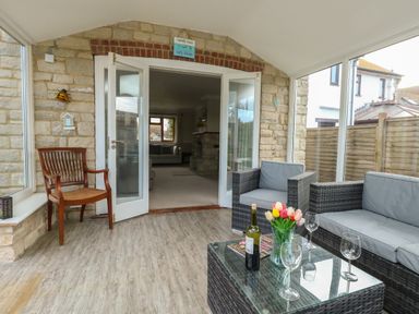 A sunroom with wicker furniture a glass coffee table with wine glasses and flowers next to a stone wall and open doors leading to a living room at Gabrianne in Burton Bradstock