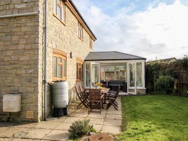 A stone house with wooden windows and a conservatory attached with glass doors opening to a patio with a wooden dining table and chairs in a garden at Gabrianne in Burton Bradstock