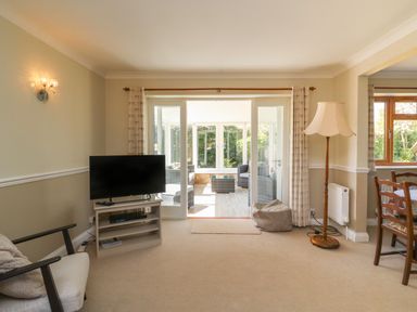 A living room with a television on a stand armchair floor lamp and sliding glass doors leading to a sunroom at Gabrianne in Burton Bradstock