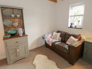 A small sitting area with a leather sofa, cushions, a quilt, a cabinet with plates and bowls, and a window with blinds at Little House in Red Wharf Bay