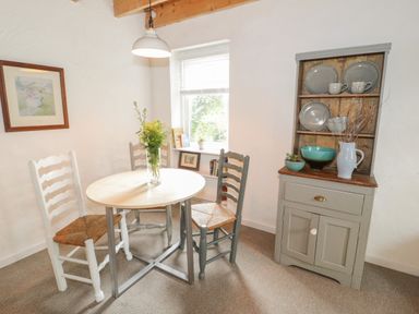 A dining area with a round table and four chairs next to a window and a wooden cabinet with dishes at Little House in Red Wharf Bay
