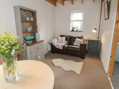 A small living room with a brown leather sofa a side table with a lamp a cabinet with dishes and a round wooden table with flowers at Little House in Red Wharf Bay
