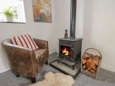 A leather armchair with a cushion beside a wood burning stove with fire and a basket of firewood at Little House in Red Wharf Bay