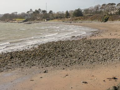 A beach with waves and rocky shoreline with trees and buildings in the background at Little House in Red Wharf Bay