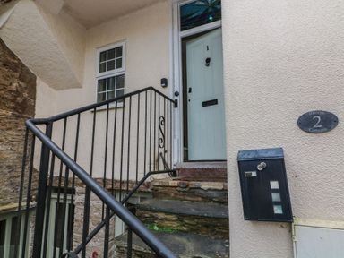 A staircase with black metal railing leading to a pale blue door with the number 1 the exterior wall has a mailbox and a plaque labeled Baileys 2 Cottages at 1 Bailey's Cottage in Stoke Fleming