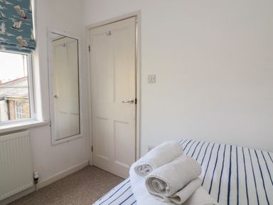 A bedroom corner with a door a wall mirror a window with a ship patterned blind and towels on a bed at 1 Bailey's Cottage in Stoke Fleming