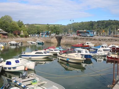 A harbor with multiple small boats docked and a stone wall with buses cars and trees in the background at 1 Bailey's Cottage in Stoke Fleming