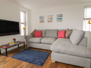 A living room with a grey sectional sofa with red and brown cushions a blue rug a wall mounted TV and small windows at Elin Howlyek in Newquay