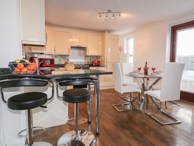 A kitchen with a black countertop bar with two black stools and a dining area with a glass table and four white chairs at Elin Howlyek in Newquay