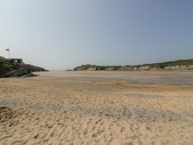A sandy beach with a small building and a flag on the left and rocky cliffs in the background at Elin Howlyek in Newquay