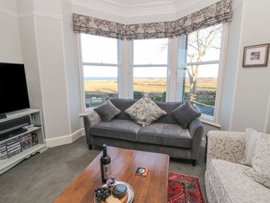 A living room with a grey sofa and a wooden coffee table in front of bay windows showing a rural view at Whinstones at 4 The Villas in Embleton