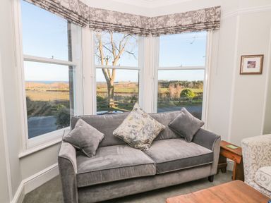 A gray sofa with cushions in front of bay windows overlooking a rural landscape at Whinstones at 4 The Villas Embleton