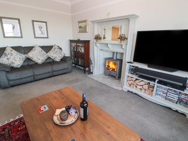 A living room with a grey sofa and floral cushions a wooden coffee table a fireplace with a wood stove and a TV on a white stand filled with wood and DVDs at Whinstones at 4 The Villas in Embleton