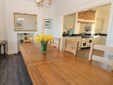 A dining room with a wooden table and chairs with yellow flowers in a vase and a kitchen with cabinets and a stove at Whinstones at 4 The Villas in Embleton