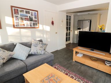 A living room with a sofa and cushions a wooden TV stand with a television and a coffee table a kitchen with a refrigerator in the background at Whinstones at 4 The Villas in Embleton