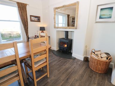 A dining room with wooden chairs and table a wood burning stove with a fire basket of logs and a large mirror on the wall at Whinstones at 4 The Villas Embleton