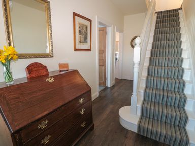 A hallway with a wooden chest of drawers with flowers on top and a staircase with striped carpet at Whinstones at 4 The Villas in Embleton