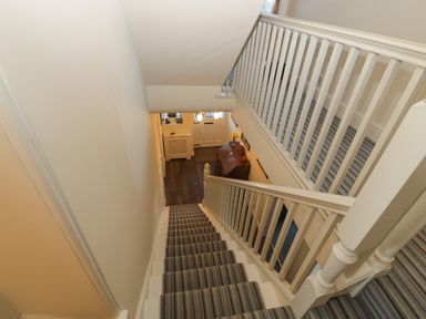 A staircase with striped carpet leading down to a hallway with a wooden floor a small cabinet and a door at Whinstones at 4 The Villas Embleton