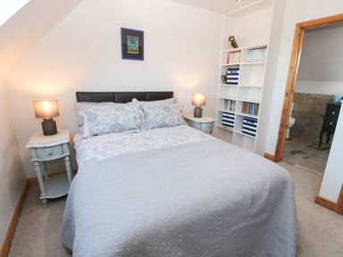 A bedroom with a bed flanked by two bedside tables with lamps and a white shelving unit next to a doorway leading to a bathroom at Whinstones at 4 The Villas in Embleton