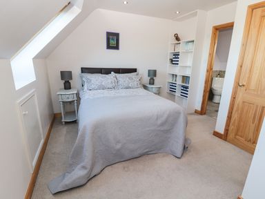 A bedroom with a bed two bedside tables with lamps a bookshelf and an open door showing a bathroom at Whinstones at 4 The Villas Embleton