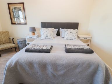 A bedroom with a bed with pillows and folded towels on a coverlet two nightstands a lamp an armchair a wicker basket and framed artwork on the wall at Whinstones at 4 The Villas in Embleton