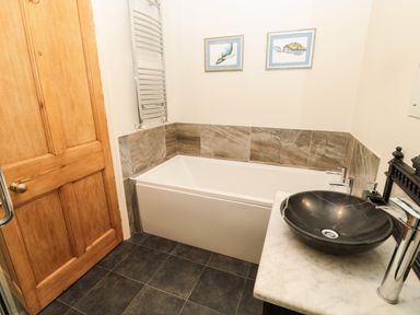 A bathroom with a bathtub, a black bowl sink on a marble countertop, framed pictures on the wall, and a wooden door at Whinstones at 4 The Villas in Embleton