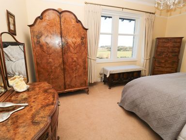 A bedroom with wooden furniture including a wardrobe dresser and chest of drawers with a window showing a garden at Whinstones at 4 The Villas Embleton