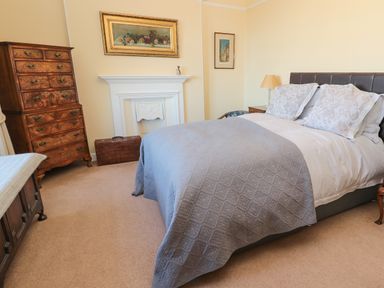 A bedroom with a bed covered in white and gray bedding a wooden chest of drawers a white fireplace and framed pictures on the walls at Whinstones at 4 The Villas in Embleton