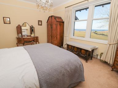 A bedroom with a bed covered by a quilt a wooden wardrobe and a dressing table with a mirror by a window at Whinstones at 4 The Villas in Embleton