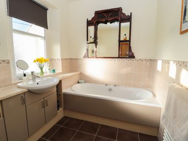A bathroom with a bathtub a sink under a window with flowers and a mirror above the bathtub at Whinstones at 4 The Villas Embleton