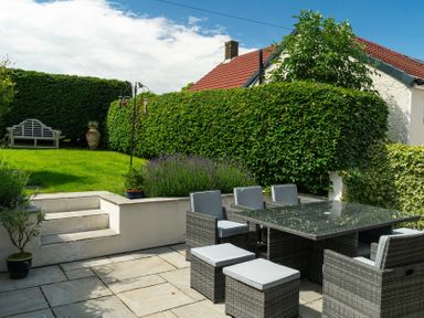 A backyard patio with a glass dining table and chairs and a grassy area with a bench and large pot at Whinstones at 4 The Villas in Embleton