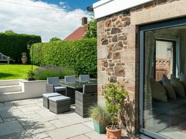 An outdoor patio with a dining table and chairs next to a stone wall and garden at Whinstones at 4 The Villas Embleton